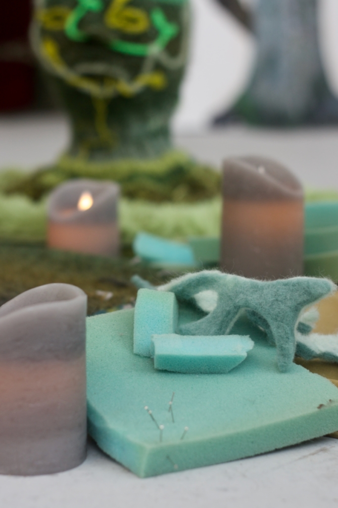 Close-up of pale green-blue craft foam, water-felted lichen-like shapes, grey LED candles, and knitted head blurry in the background