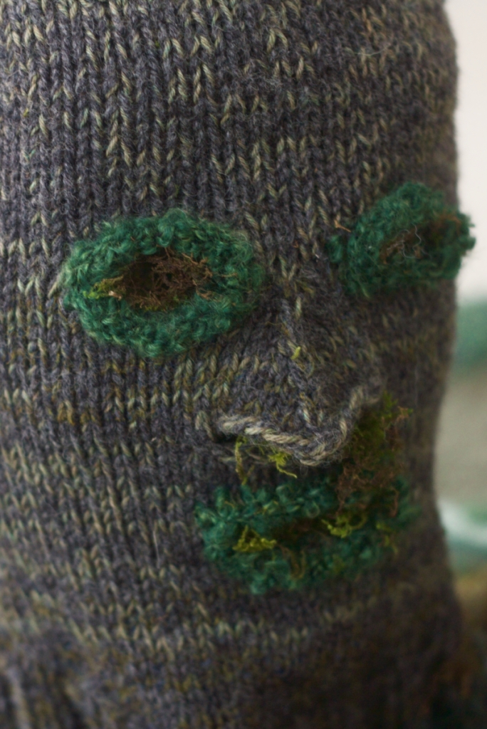 Closeup of knitted grey-green head with bright green eye and mouth openings, and real moss stuffed inside
